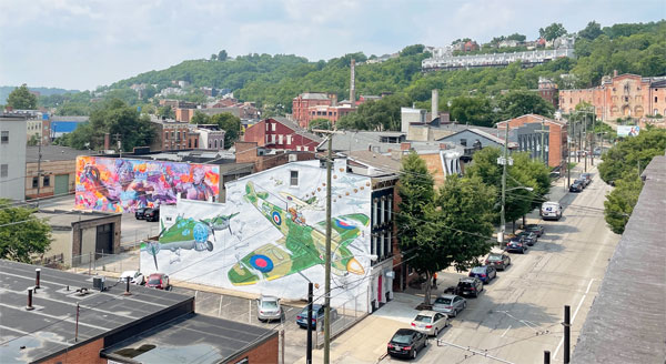 Tree covered hills lead to old brick buildings decorated with colorful murals.