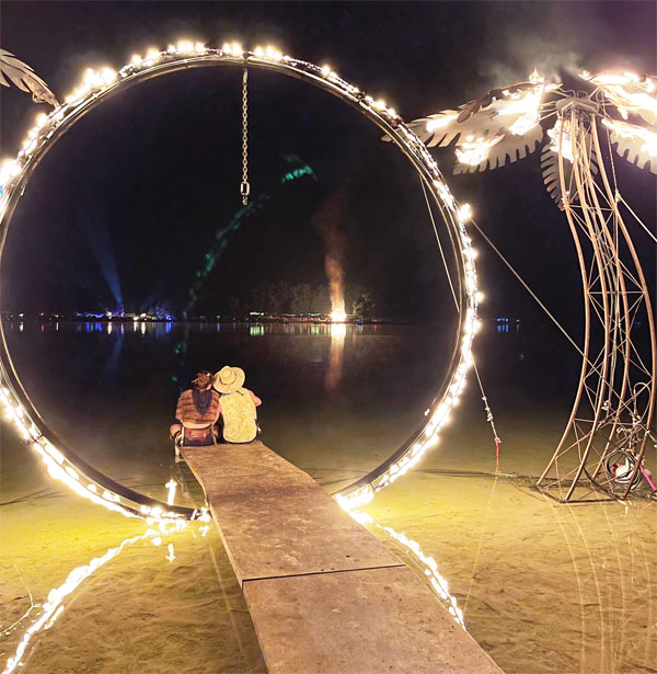 A couple sit under a flaming metal ring with metal flaming palm trees on either side.