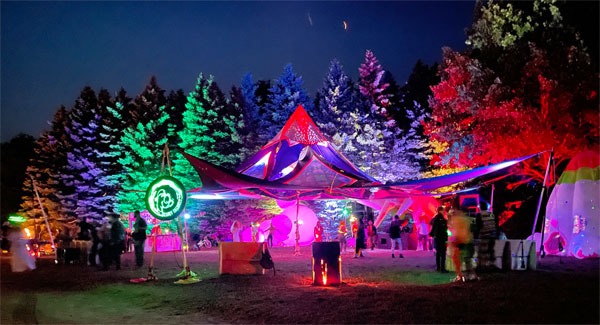 Colorful lights illuminate pine trees and a dance floor under a tent.