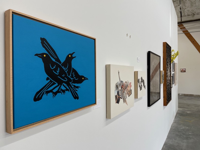 A blue and black painting of three grackles hangs on a gallery wall.