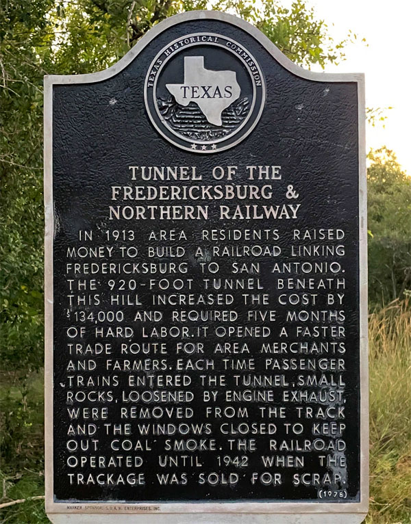 Historical sign that reads: "In 1913 area residents raised money to build a railroad linking Fredericksburg to San Antonio. The 920-foot tunnel beneath this hill increased the cost by $134,000 and required five months of hard labor. It opened a faster trade route for area merchants and farmers. Each time passenger trains entered the tunnel, small rocks, loosened by engine exhaust, were removed from the track and the windows closed to keep out coal smoke. The railroad operated until 1942 when the trackage was sold for scrap."