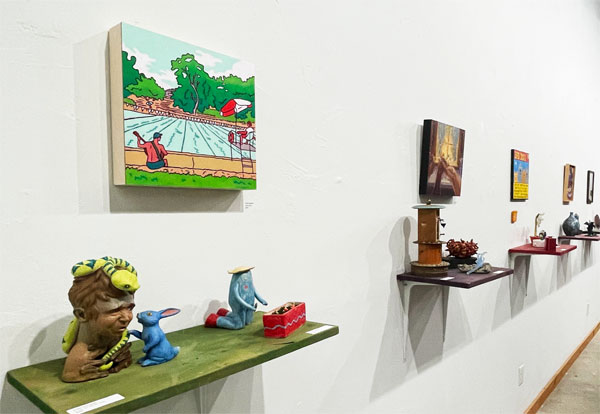Paintings and shelves with small sculptures hanging on gallery wall.